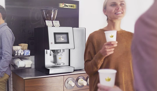 Mehrere Leute stehen im Büro neben einer Jacobs Professional Barista One PowerPack Kaffeemaschine und trinken eine Tasse Kaffee.