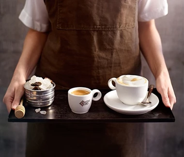 Abbildung eines Tabletts mit drei Tassen Kaffee, das von einer Frau mit brauner Schürze getragen wird.