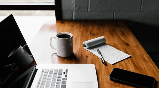 Auf einem Schreibtisch stehen Laptop und Notizblock neben einer Kaffeetasse.