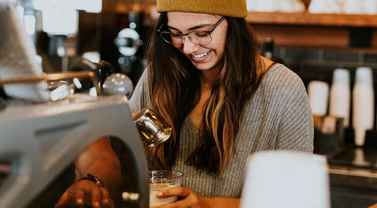 Abbildung einer jungen Barista, die lächelnd einen Kaffee zubereitet.