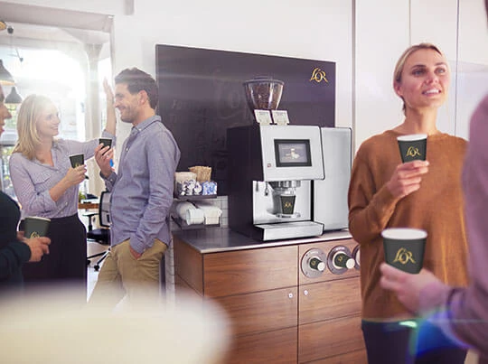 Eine L'OR Promesso Kaffeemaschine steht im Büro. Mehrere Mitarbeiter stehen daneben und trinken gerade Kaffee.