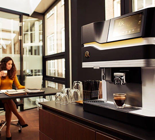 Abbildung einer L'OR Promesso Kaffeemaschine. Daneben sitzt eine Frau, die gerade eine Tasse Kaffee trinkt.