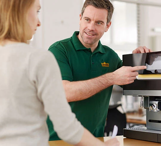 Ein Servicemitarbeiter berät gerade eine Kundin. Er steht neben einer L'OR Promesso Kaffeemaschine.