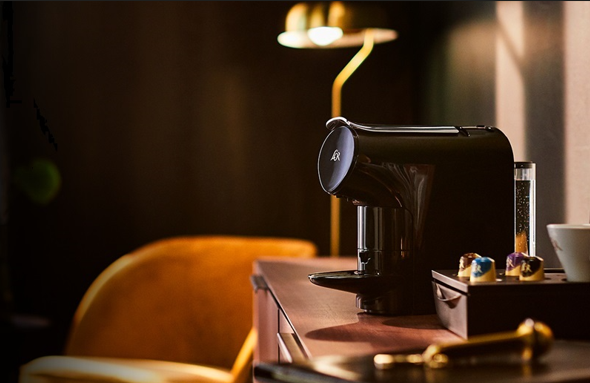 LOR Mini Kaffeemaschine auf Holzoberfläche mit Kapselbehälter; im Hintergrund gelber Sessel und Lampe.