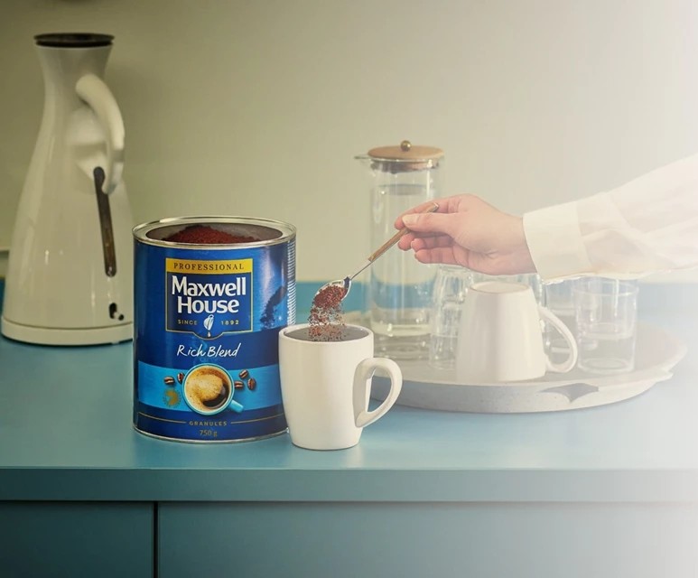 Hand mit Löffel voller Kaffeekörnchen über weißer Tasse, daneben geöffnete Dose Maxwell House Professional Rich Blend.