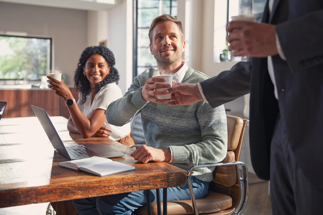Drei Personen in einem modernen Büro bei einem Meeting, eine reicht der anderen eine Tasse.