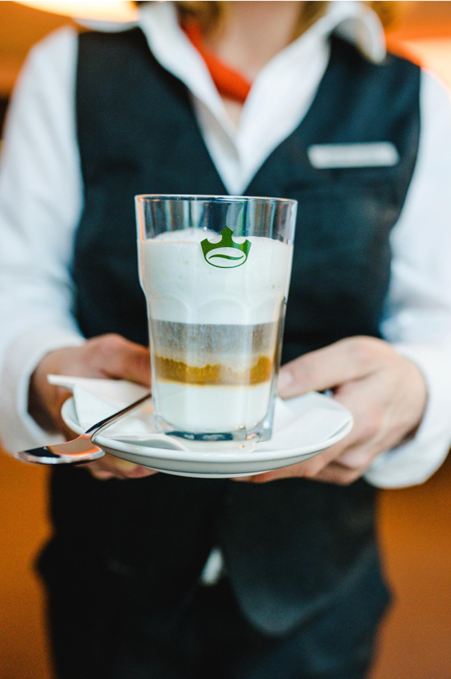 Hotelmitarbeiterin serviert  Latte Macchiato im Jacobs Glas